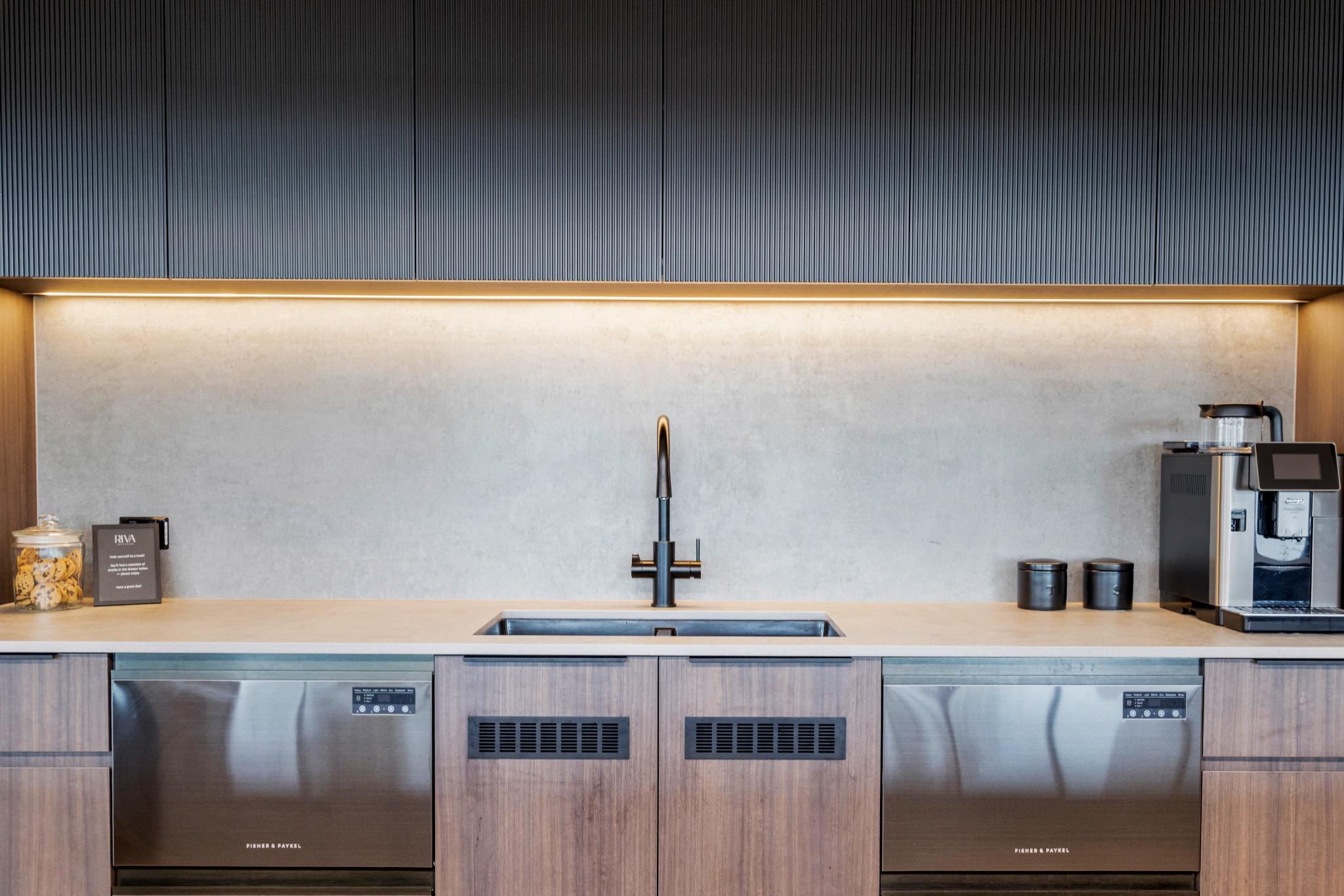 Compact office kitchenette featuring black tapware, stone benchtop, and overhead cabinetry with integrated lighting.