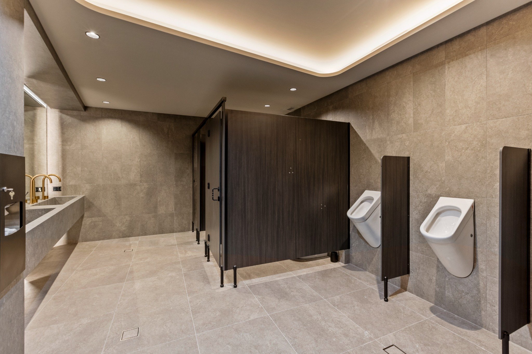Premium commercial male amenities at Riva Robina featuring full-height stone-look tiles, matte black toilet partitions, floating concrete vanity, brushed brass sensor tapware, and concealed lighting details.