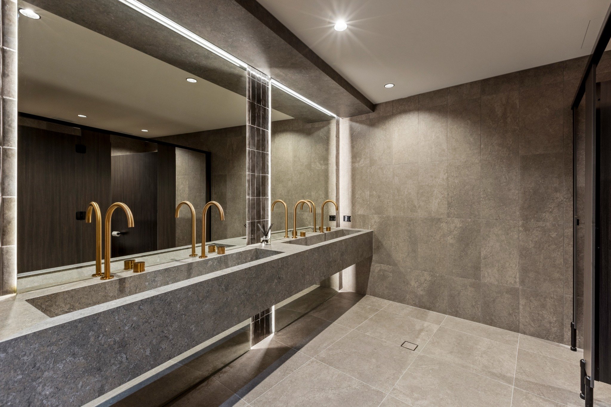 Detail view of the men’s washroom at Riva Robina with floating stone vanity, integrated LED mirror lighting, brushed gold tapware, and large-format grey tiles for a refined, modern finish.