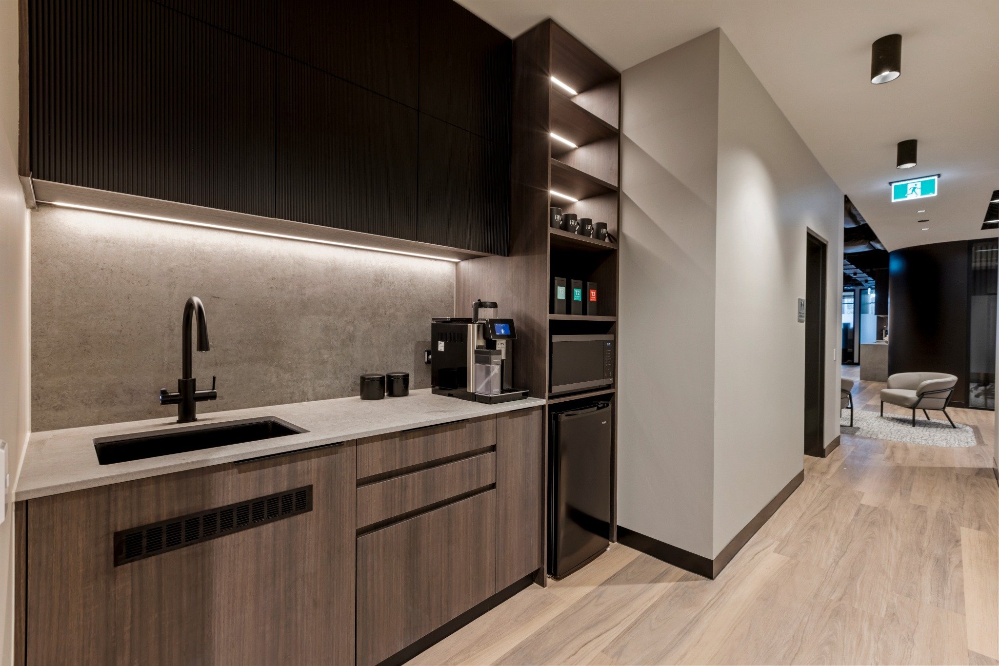 Compact staff kitchenette at Riva Robina featuring matte black tapware, textured upper cabinets, full-height open shelving, and integrated under-bench fridge and microwave in a warm timber palette.