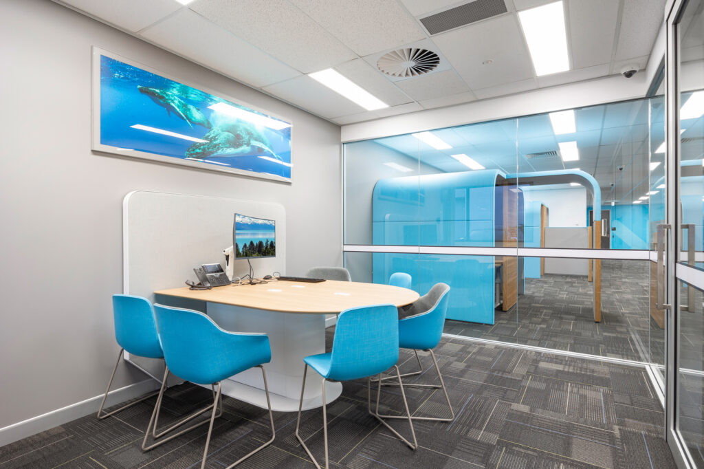 Modern office workspace featuring a round table with blue chairs, a computer setup, and a large aquatic-themed wall art, designed for collaborative environments in commercial interiors.