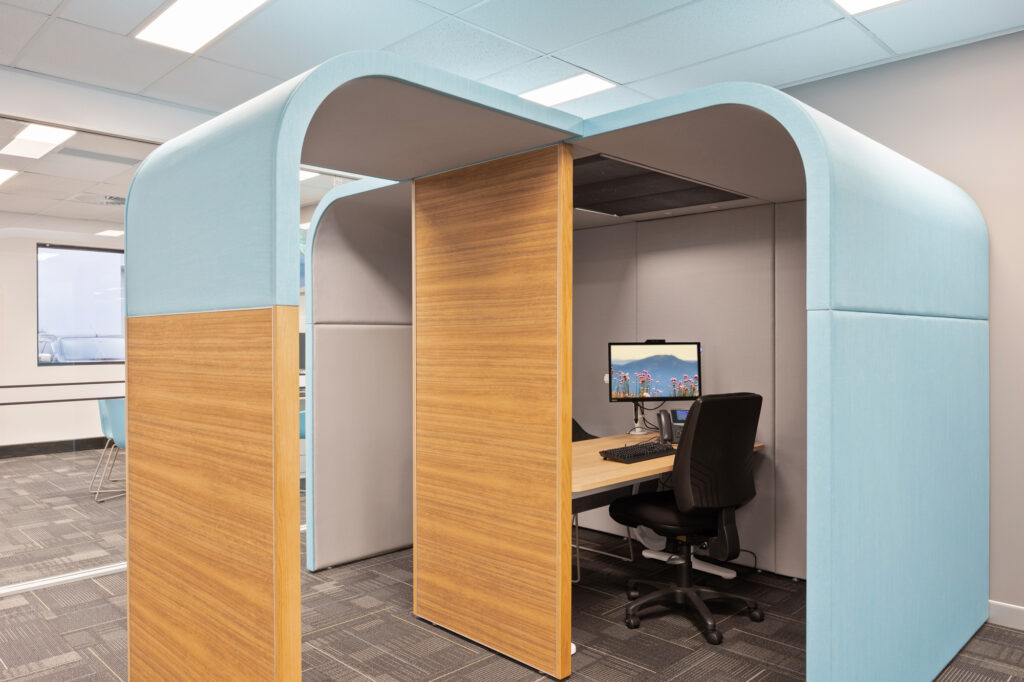Modern office workspace design featuring a private workstation with a wooden desk, ergonomic chair, and computer, enclosed by sound-absorbing panels in blue and grey, reflecting contemporary commercial interior design principles.