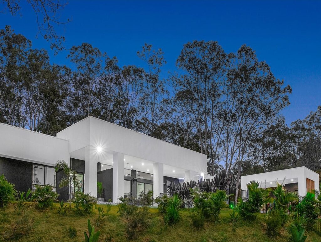 Modern residential home design in Pullenvale, Queensland, featuring sleek white architecture, large windows, and surrounding greenery at dusk.