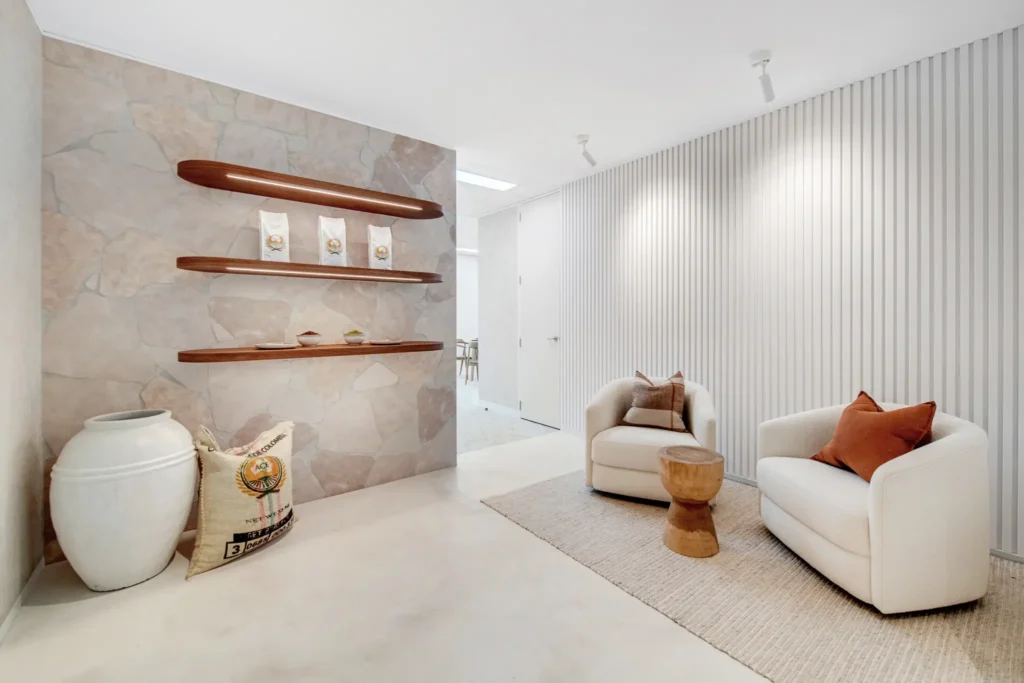 Interior design of a modern waiting area in a healthcare clinic, featuring a textured stone wall, wooden shelves displaying decorative items, white armchairs with orange cushions, and a neutral-toned rug.