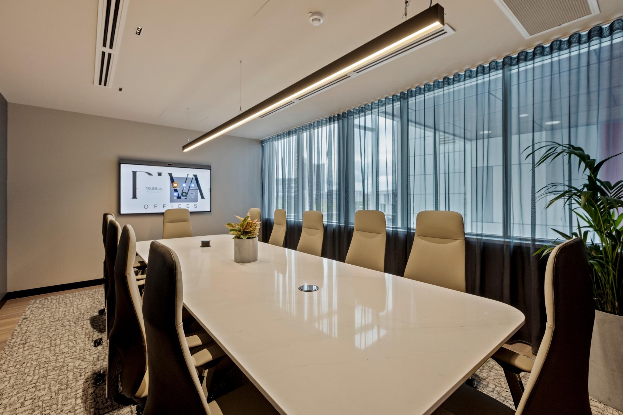 Modern conference room with elegant design, featuring a long white table, beige ergonomic chairs, large windows with sheer curtains, and a digital display showing "RVA OFFICES."