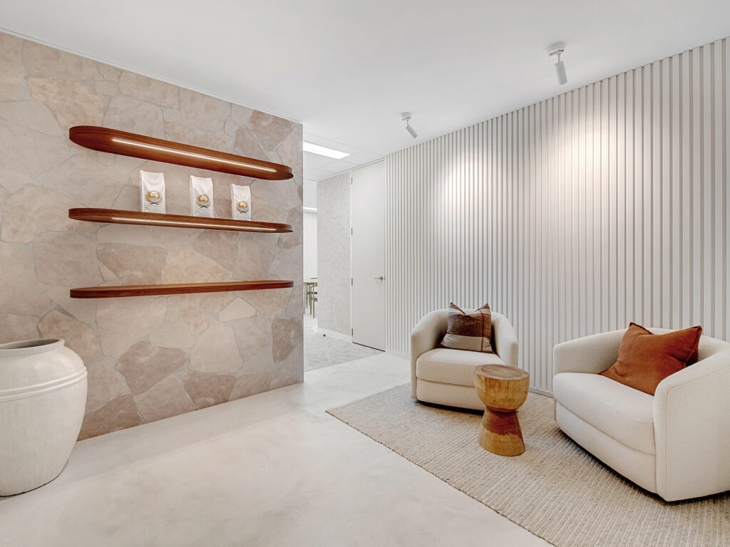 Modern commercial interior featuring textured stone wall, wooden shelves displaying products, two stylish armchairs with cushions, and a wooden side table, emphasizing architectural design and luxury fit-out by Leithal Designs.